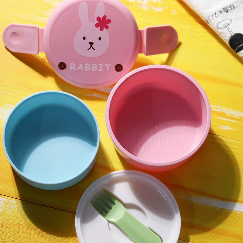 Children's kawaii food containers with pink and blue bowls, a green spoon, and a rabbit-themed lid on a yellow surface.