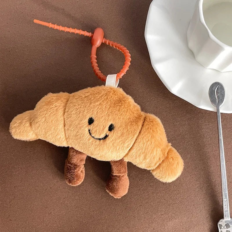 Plush kawaii toy resembling a croissant with a smiley face on a brown surface next to a white cup and saucer.