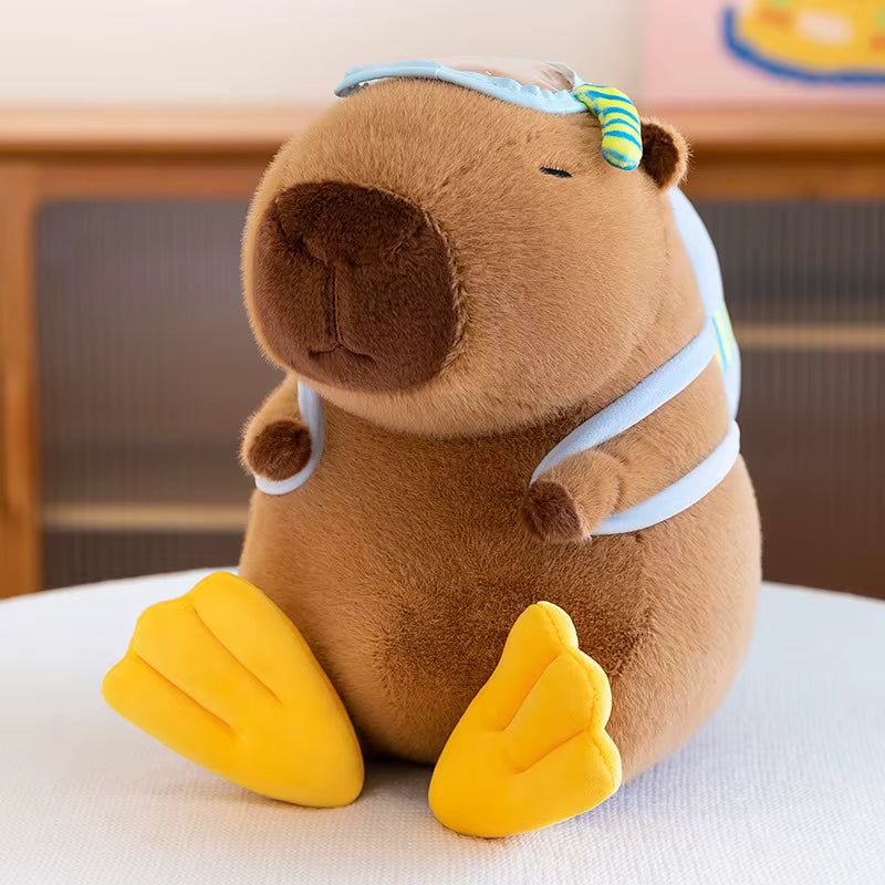 A brown capybara plush toy wearing a light blue swim cap and a green striped swim tail, sitting on a white surface with a blurred indoor background.