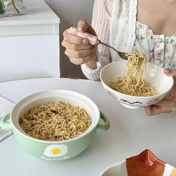 Person eating noodles from a kawaii ramen bowl with a design on a white table.