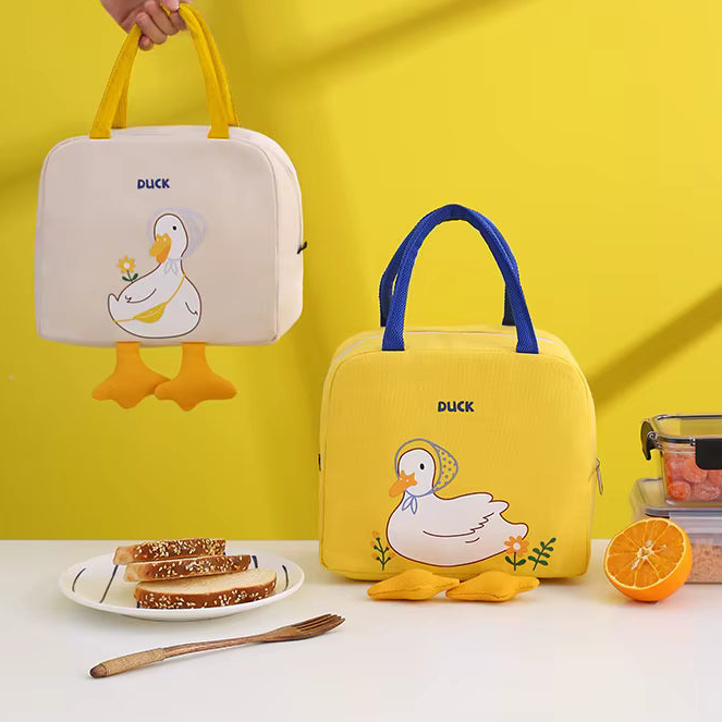 Two duck-themed lunch bags are displayed on a white tabletop against a bright yellow wall. One bag is cream with yellow handles and a yellow duck illustration, while the other is yellow with blue handles and a white duck illustration.
