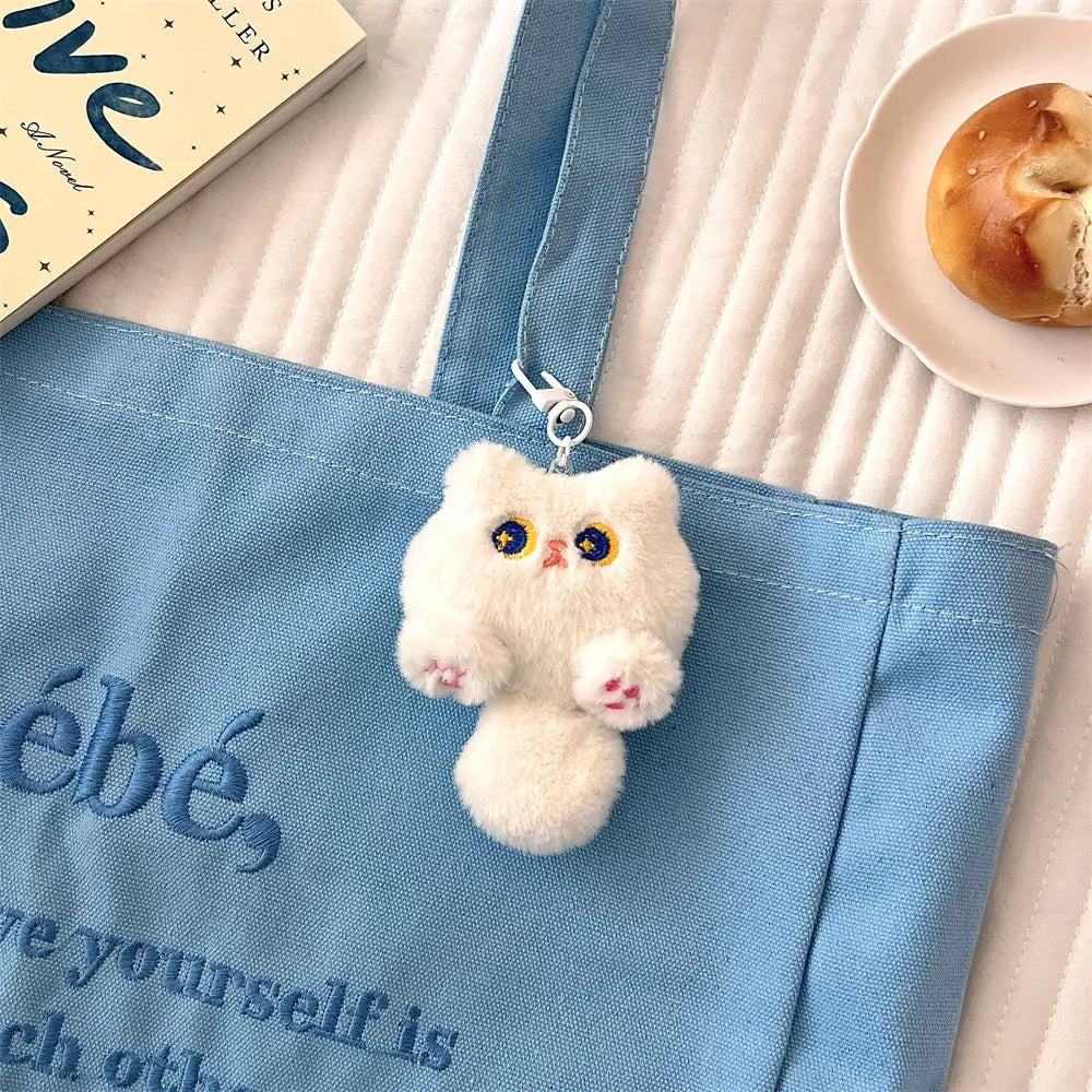 White kawaii fluffy cat-shaped keychain hanging from a blue bag with a book and pastry in the background.