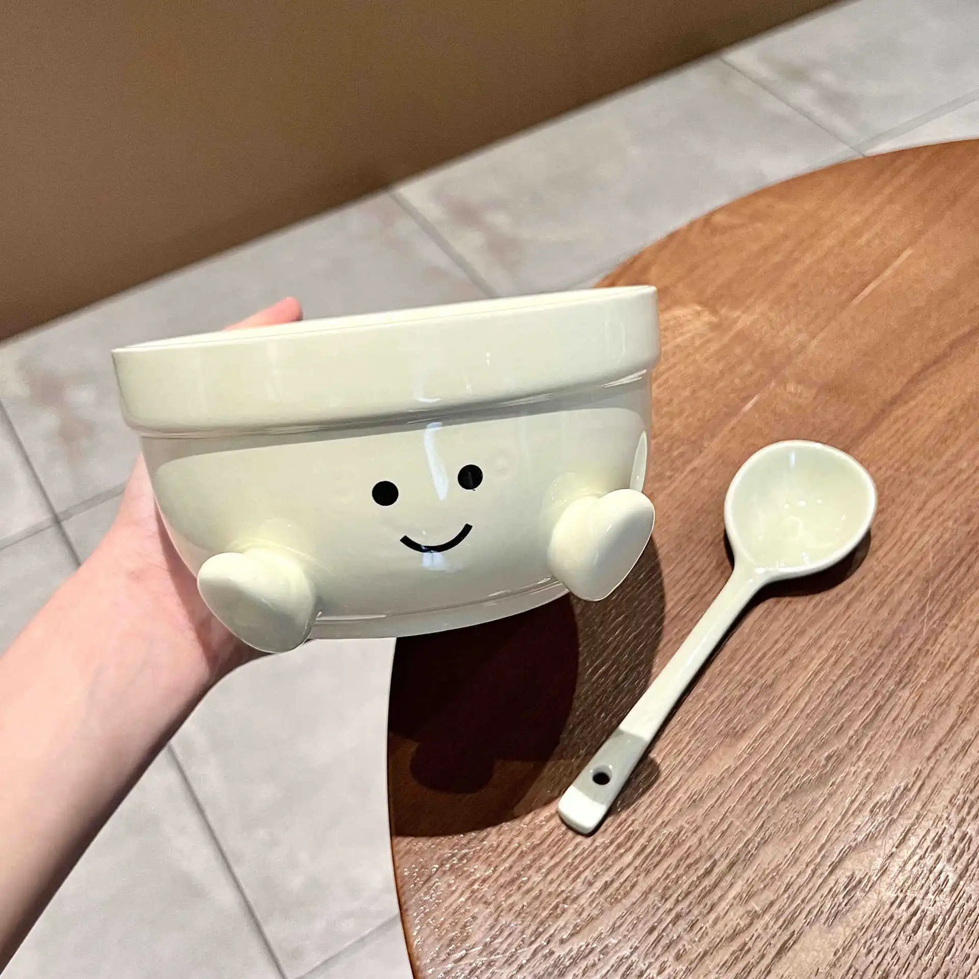 White kawaii bowl with a smiley face design held by a hand, next to a white spoon on a wooden surface.