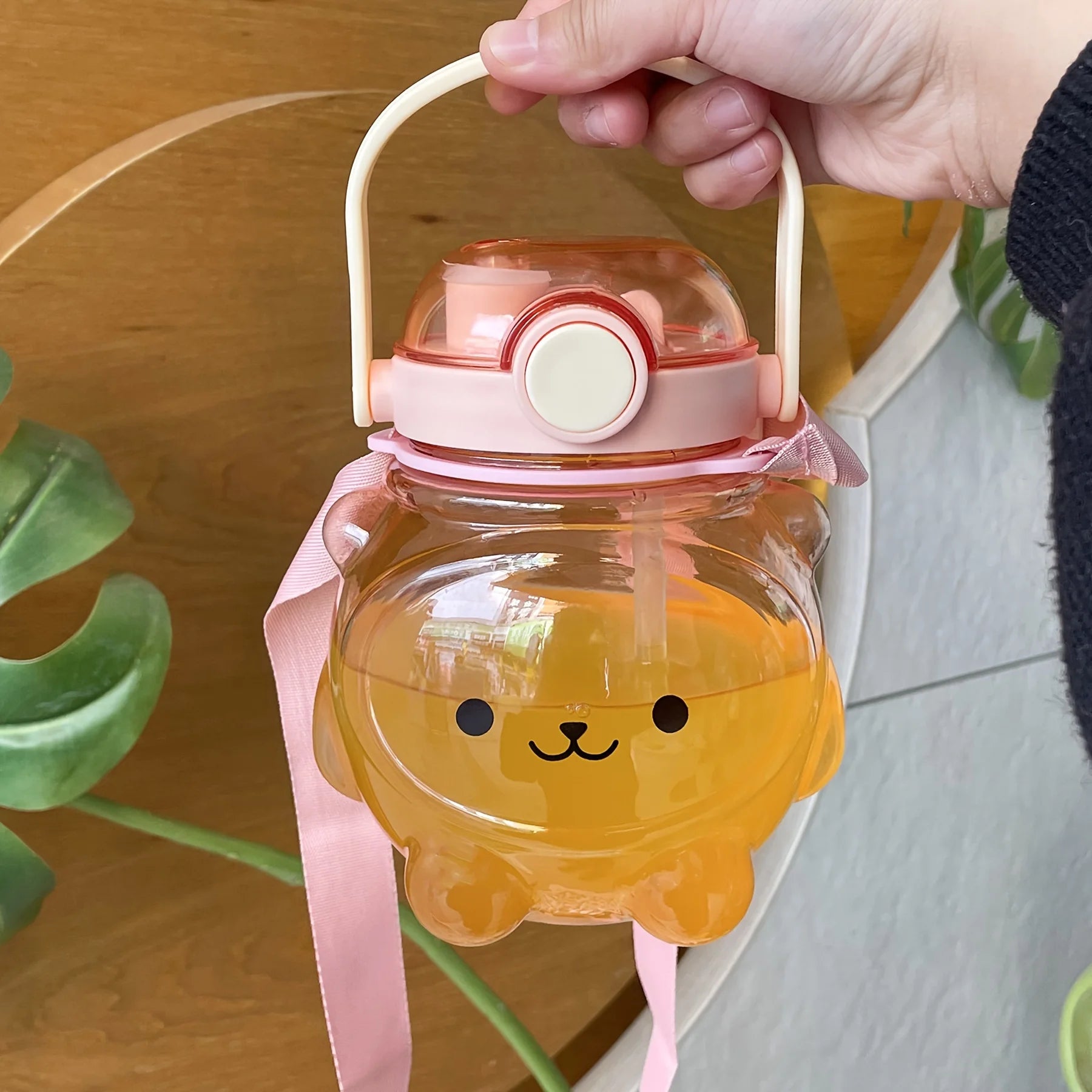 Hand holding a transparent kawaii water bottle with a bear design and pink handle and ribbon.