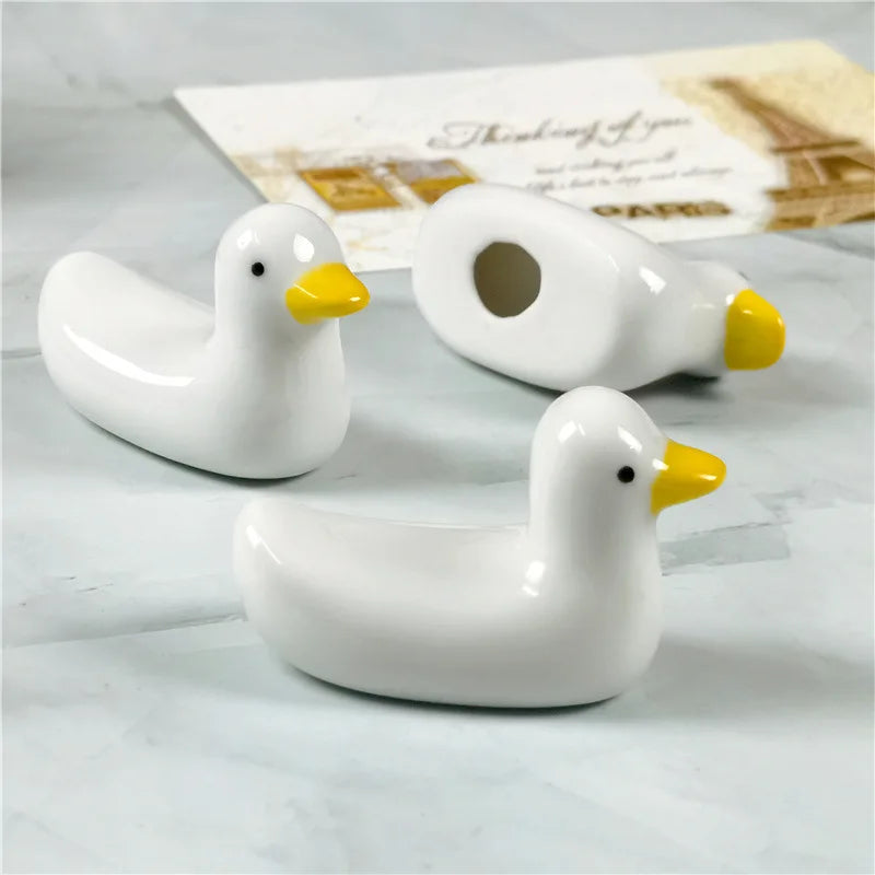 Three white ceramic ducks kawaii chopstick holders with yellow beaks on a light gray surface.
