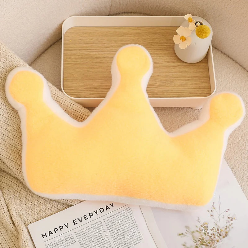 Yellow crown-shaped kawaii pillow on a beige surface with a wooden tray and decorative items.
