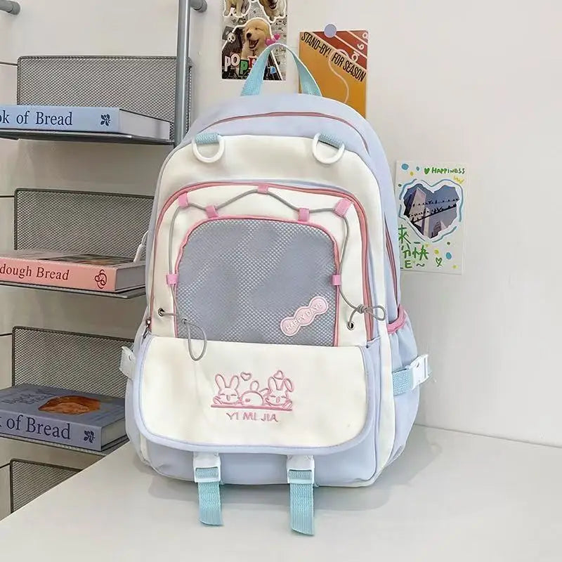 A pastel blue and white school backpack with pink accents, featuring a front mesh pocket and a white front flap with a bunny graphic and the text 'YI MEI JIA'.