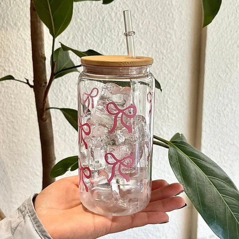 Clear glass kawaii cup with pink bow designs, wooden lid, and straw held by a hand with green leaves in the background.