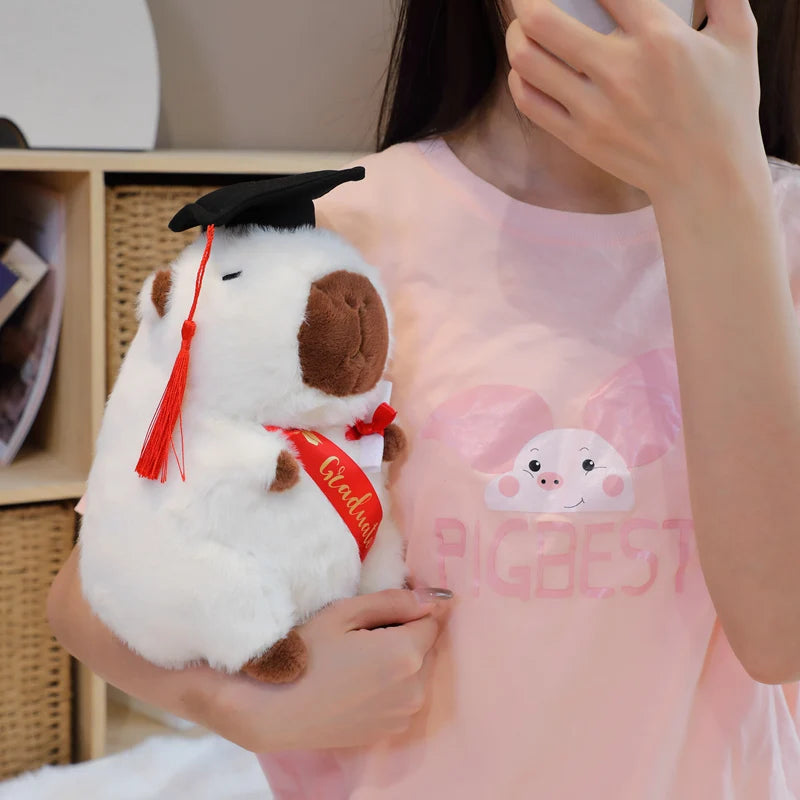 Person holding a capybara plush toy with a graduation cap and red ribbon, wearing a pink dress with a cartoon pig design.