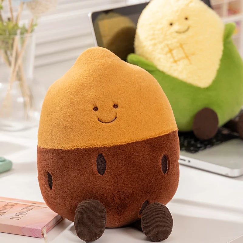 A cute acorn-shaped plush toy with a smiling face, sitting on a white desk next to a book, with a laptop and another plush toy in the background.