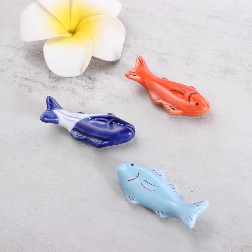 Three fish-shaped kawaii chopsticks holders in blue, orange, and white on a light gray surface with a white flower in the background.