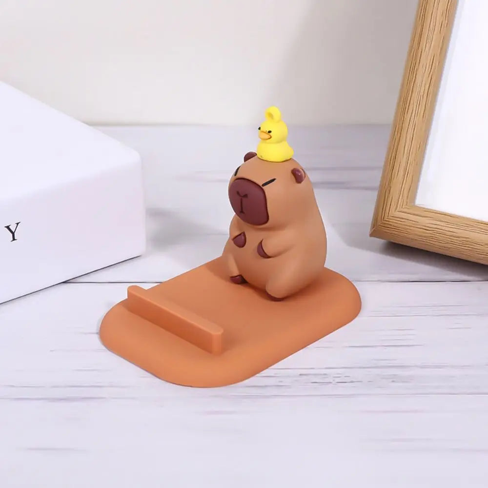 Cute capybara-themed kawaii phone stand with a small yellow duck on a brown bear's head, placed on a light wooden surface.