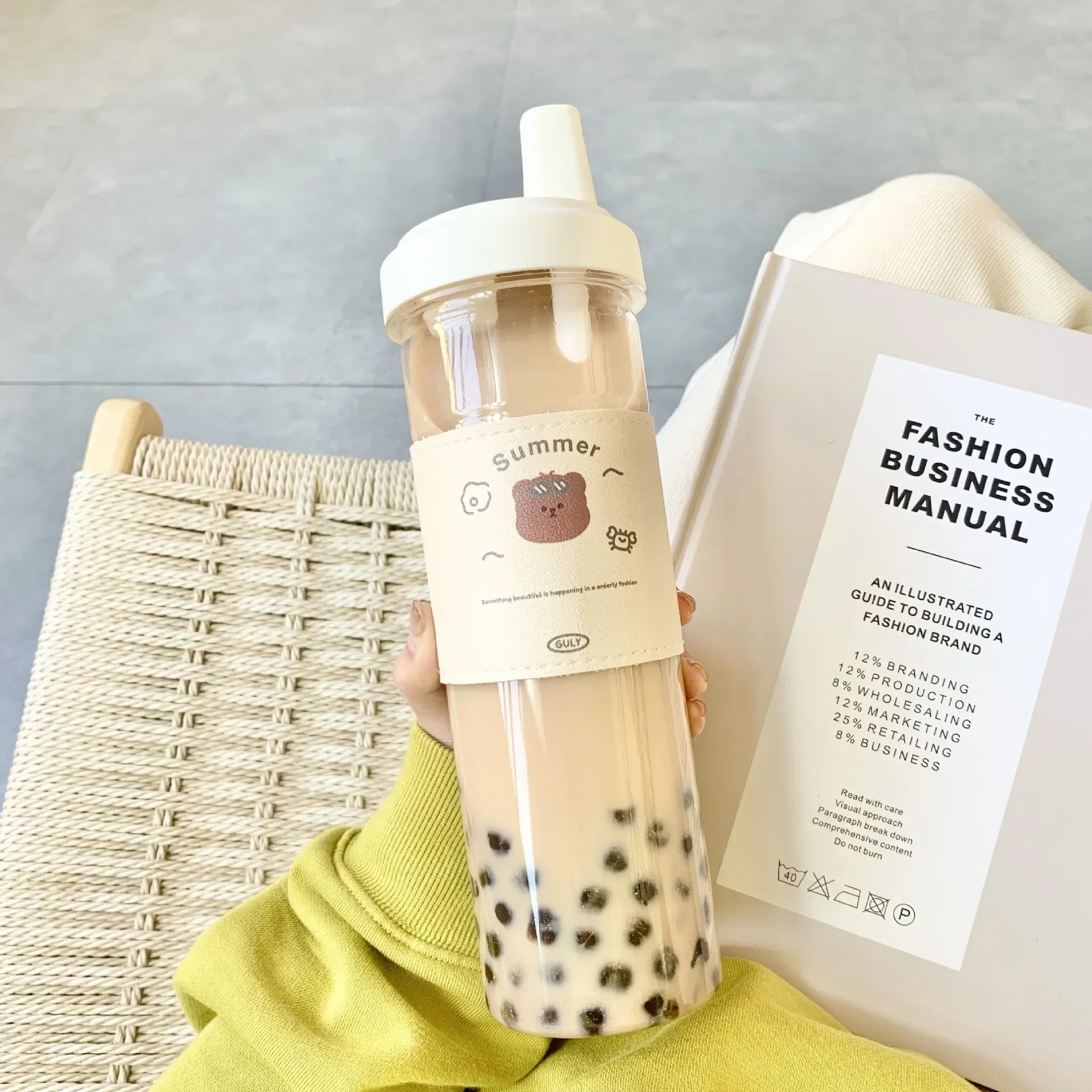 Person holding a kawaii water bottle and a book titled 'The Fashion Business Manual' on a light gray background.