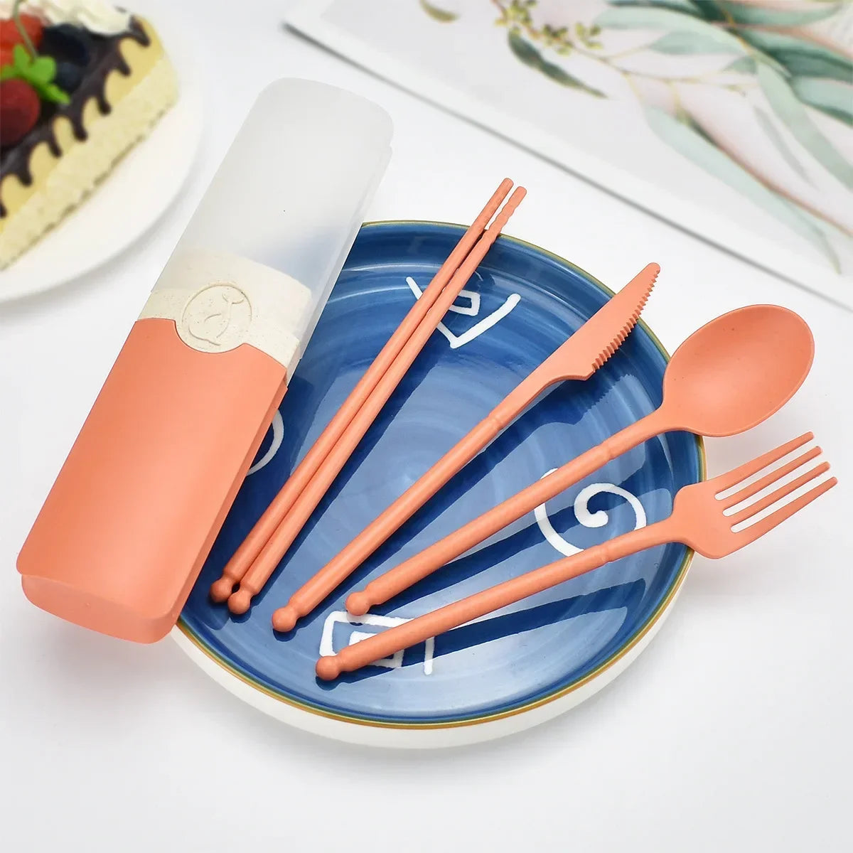 Set of pink kawaii utensils with a case on a blue plate, with a cake in the background.