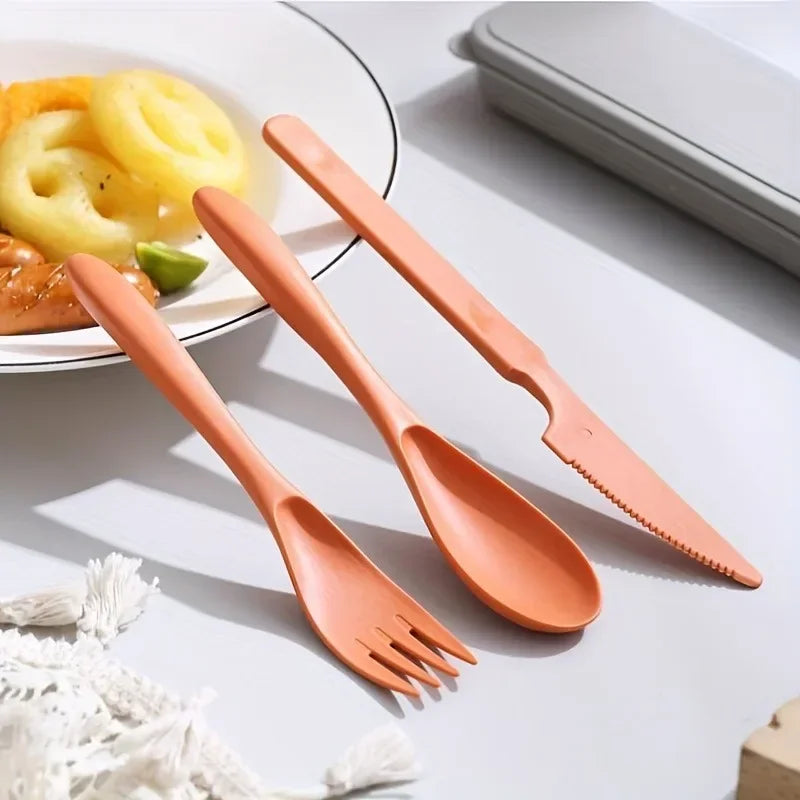Set of orange kawaii utensils including a fork, spoon, and knife on a white surface with a plate of food in the background.