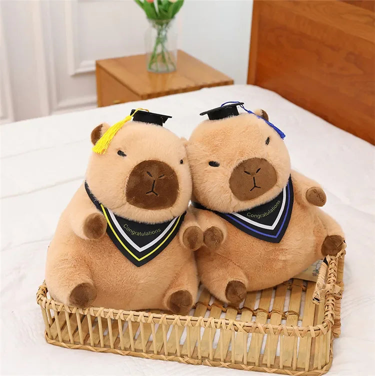Two brown kawaii plush animals with graduation caps and gowns sitting in a woven basket on a bed.