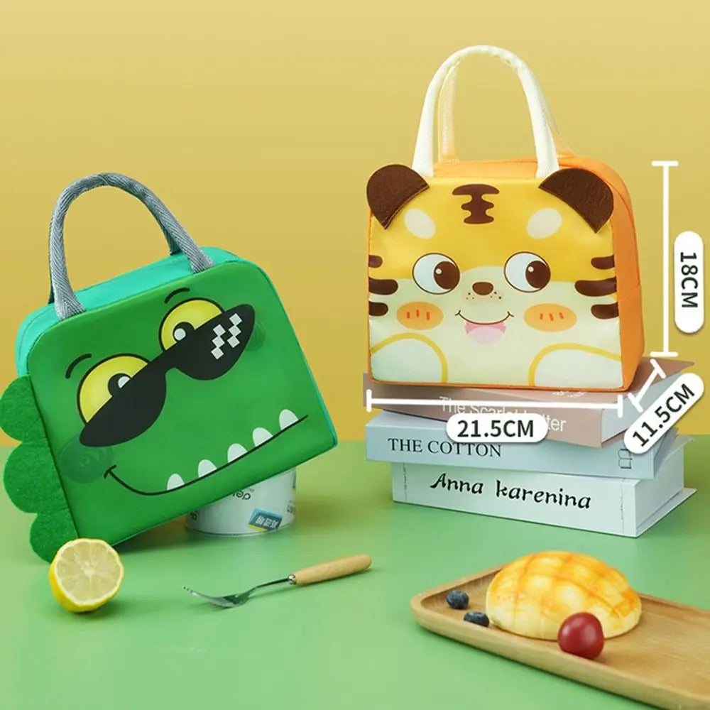 Two cartoon-themed kawaii lunch bags, one green with a dinosaur design and one yellow with tiger ears, on a green surface with books and food items.