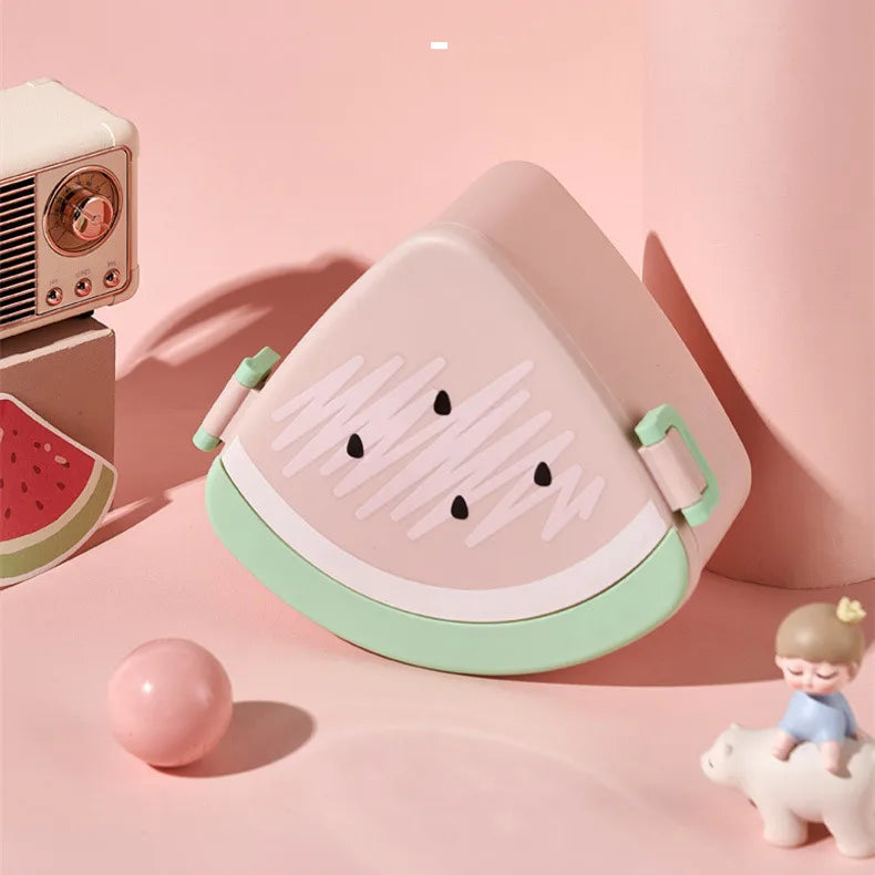 Pink and green watermelon-shaped kawaii bento box on a pink background with small decorative items.