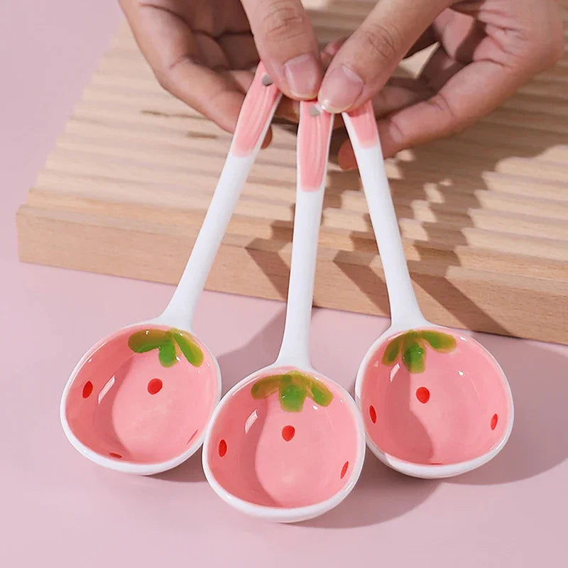 Three strawberry-themed kawaii spoons held by a hand on a pink background