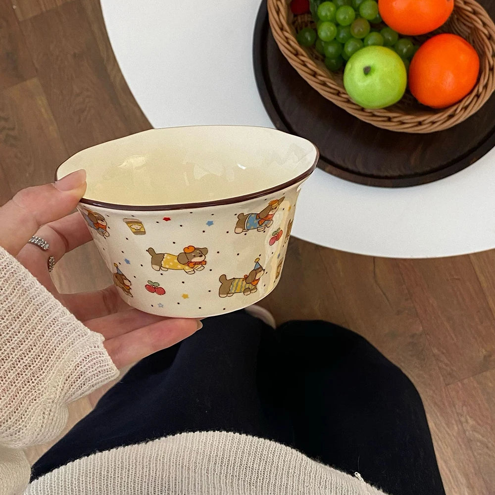 Person holding a decorative kawaii ramen bowl with cartoon characters, next to a basket of fruits on a wooden floor.
