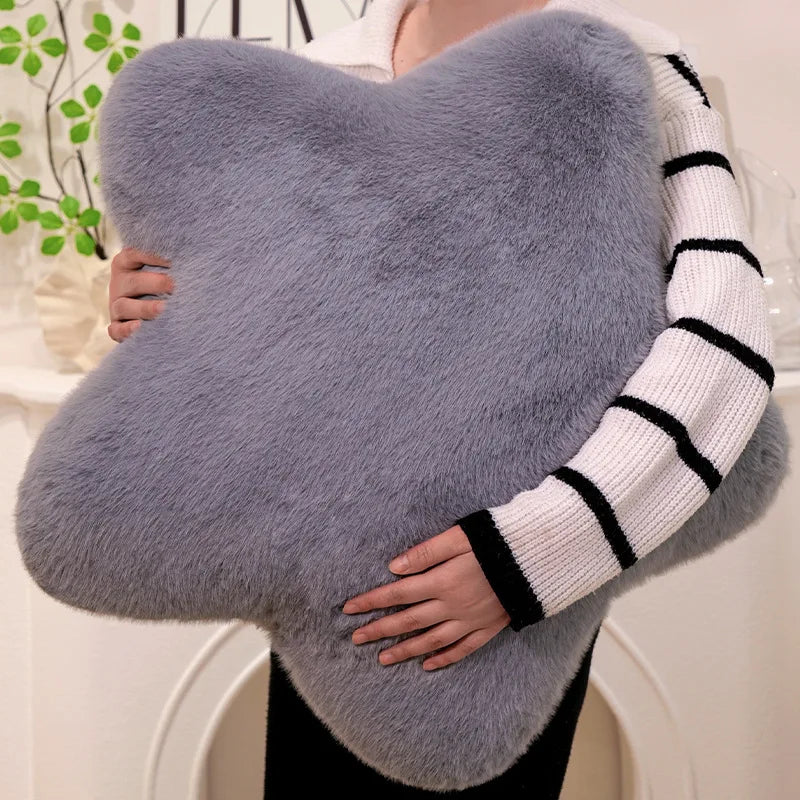 A person holding a large, fluffy, star-shaped gray cushion or pillow. The person is wearing a white knit sweater with black stripes on the sleeve, set against a soft, light-colored indoor background with a blurred green plant.