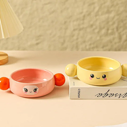Two cute ceramic kawaii ramen bowls, one pink and one yellow, with face designs on a beige surface.