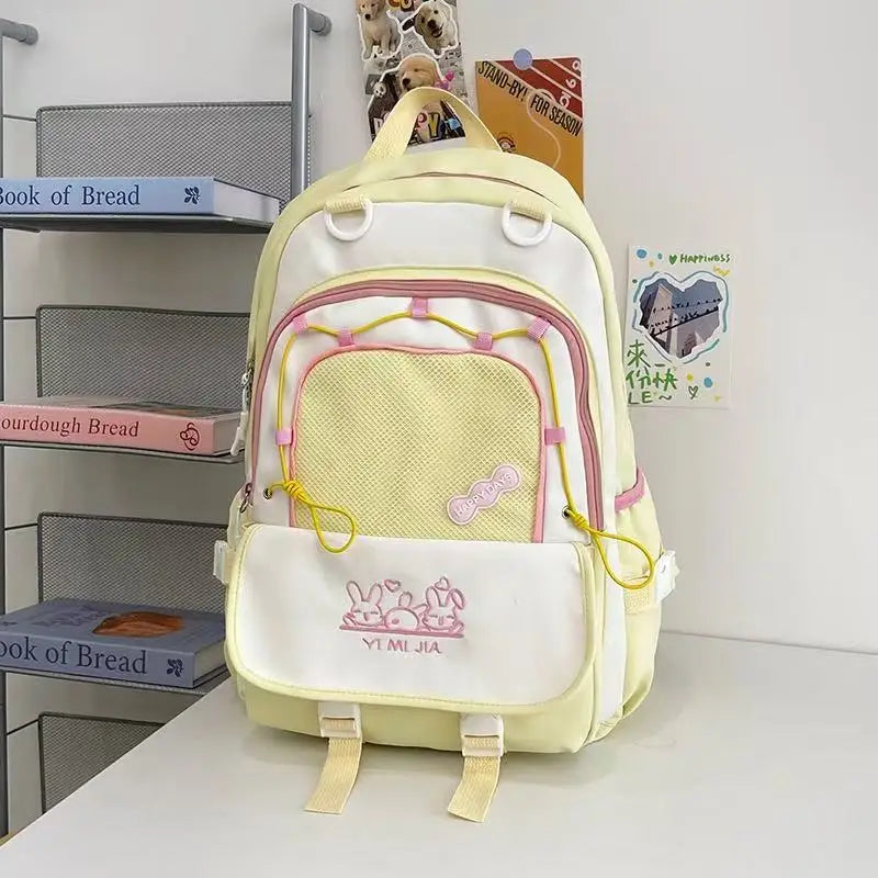 A pastel yellow and white school backpack with pink zipper accents and cartoon bunny graphics on the front pocket, displayed on a white table against a white wall with decorative books and cards.