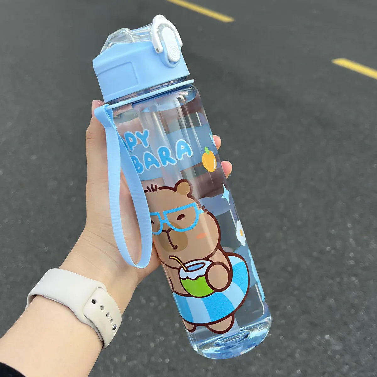 Cartoon bear kawaii water bottle with blue lid and strap held by a hand on a dark surface