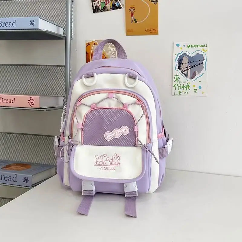 A pastel purple and white school backpack with pink zipper accents and cartoon bunny graphics on the front pocket, displayed on a white table against a white wall with decorative books and cards.