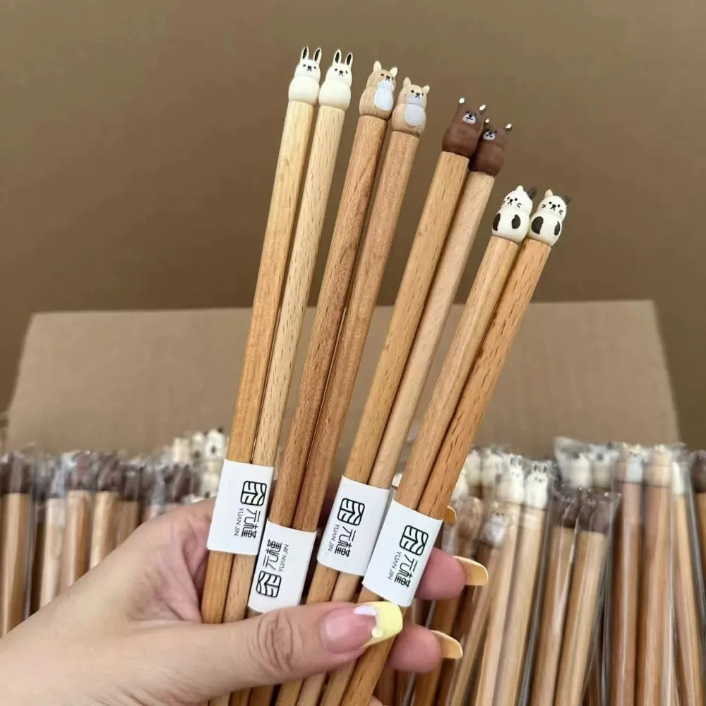 Hand holding a set of wooden kawaii chopsticks with animal-shaped tips against a cardboard box background.
