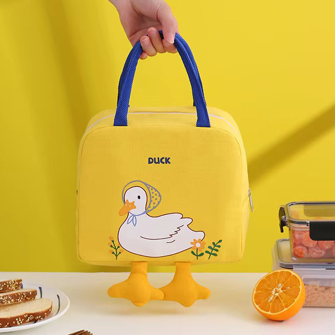A yellow insulated lunch bag with blue handles, featuring a cartoon duck character and the word 'DUCK' on the front. A hand is holding the bag, and it is styled on a white table with food props against a bright yellow background.