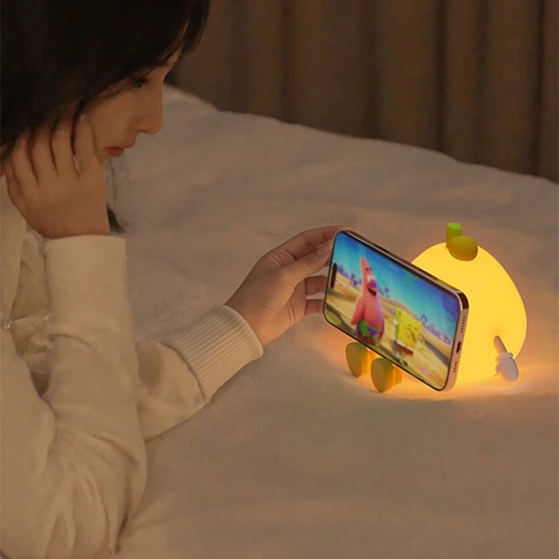 Person watching a cartoon on a smartphone with a glowing kawaii night light on a bed.