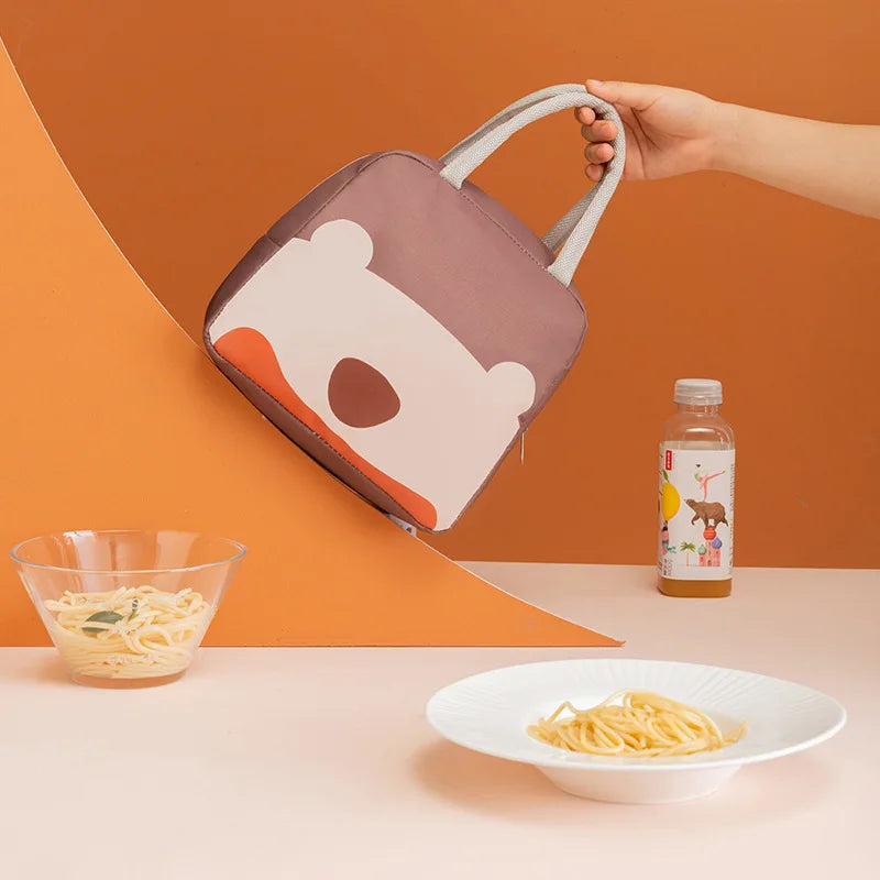 Hand holding a cartoon-themed kawaii lunch bag with food and a drink on a table against an orange background
