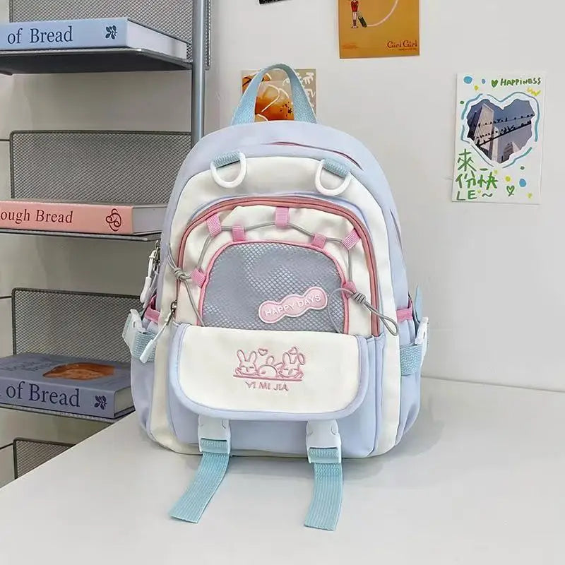 A pastel purple and white school backpack with pink zipper accents and cartoon bunny graphics on the front pocket, displayed on a white table against a white wall with decorative books and cards.