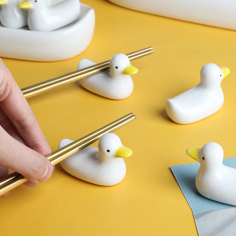 White duck-shaped kawaii chopstick holders on a yellow surface with a hand holding gold chopsticks.