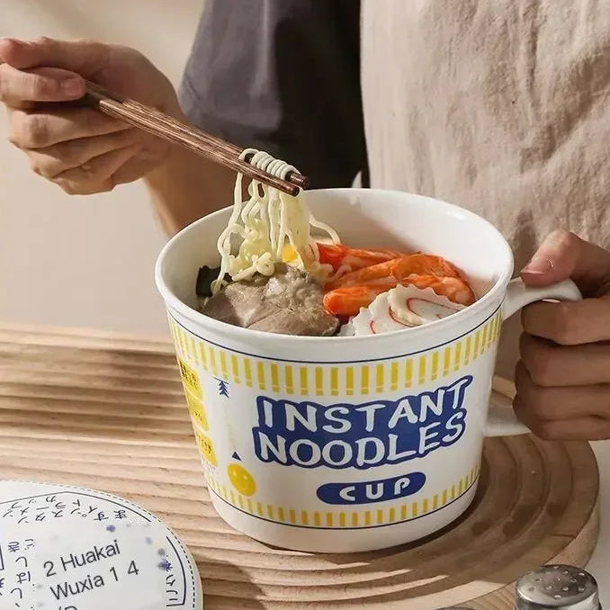 Person holding a kawaii ramen bowl of instant noodles with chopsticks on a wooden surface.