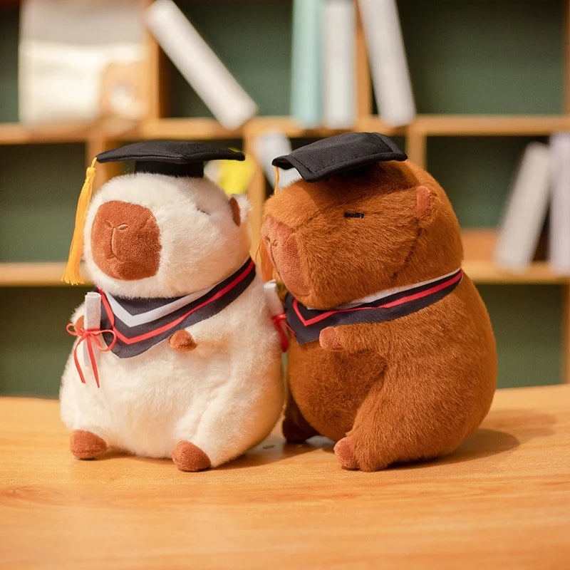 Two capybara plush toys, one white and one brown, wearing graduation caps and gowns on a wooden surface.