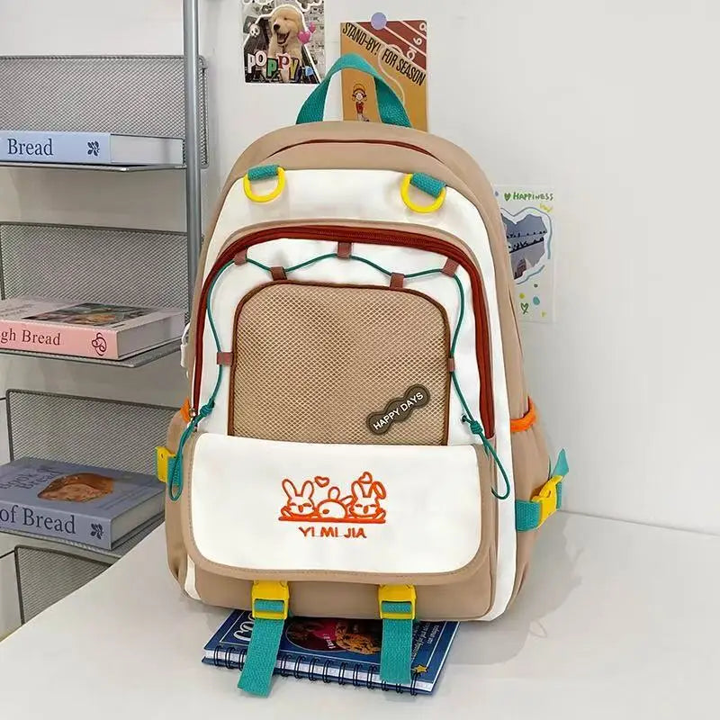 A pastel brown and white school backpack with pink zipper accents and cartoon bunny graphics on the front pocket, displayed on a white table against a white wall with decorative books and cards.