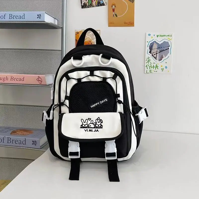 A pastel black and white school backpack with pink zipper accents and cartoon bunny graphics on the front pocket, displayed on a white table against a white wall with decorative books and cards.