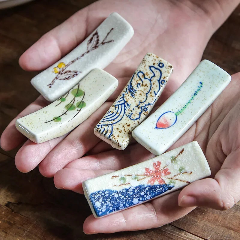 Hand holding five small ceramic kawaii chopstick holders with floral and abstract designs on a wooden surface.