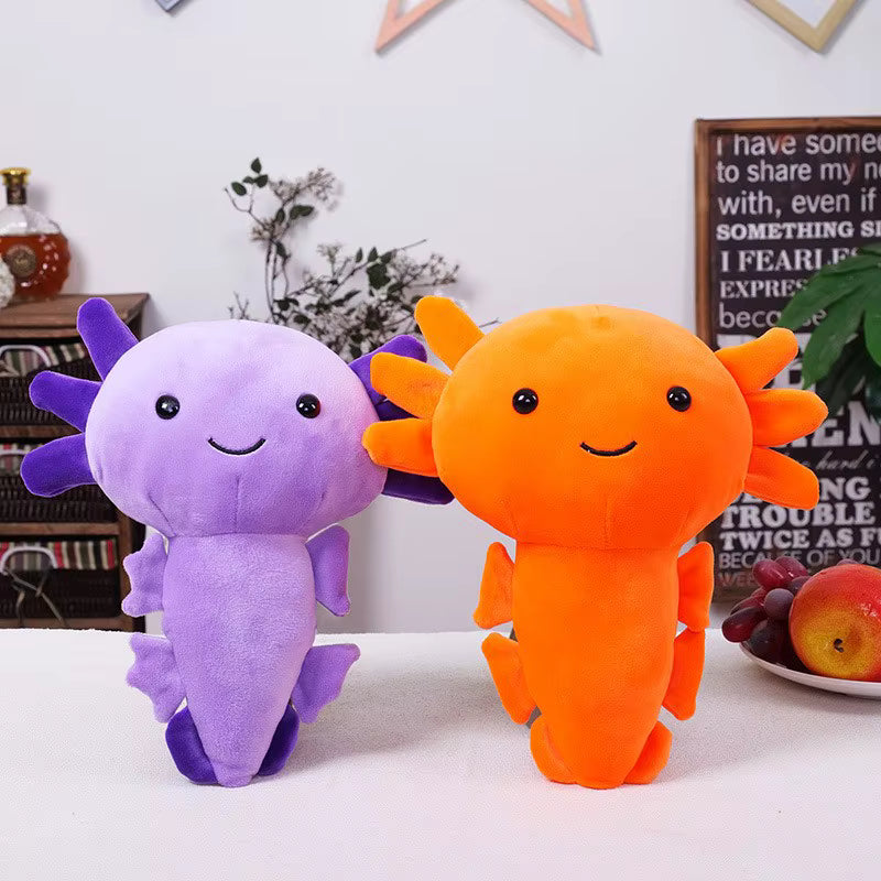 Two cute plush axolotl toys, one purple and one orange, sitting side-by-side on a white table with a cozy, styled background.
