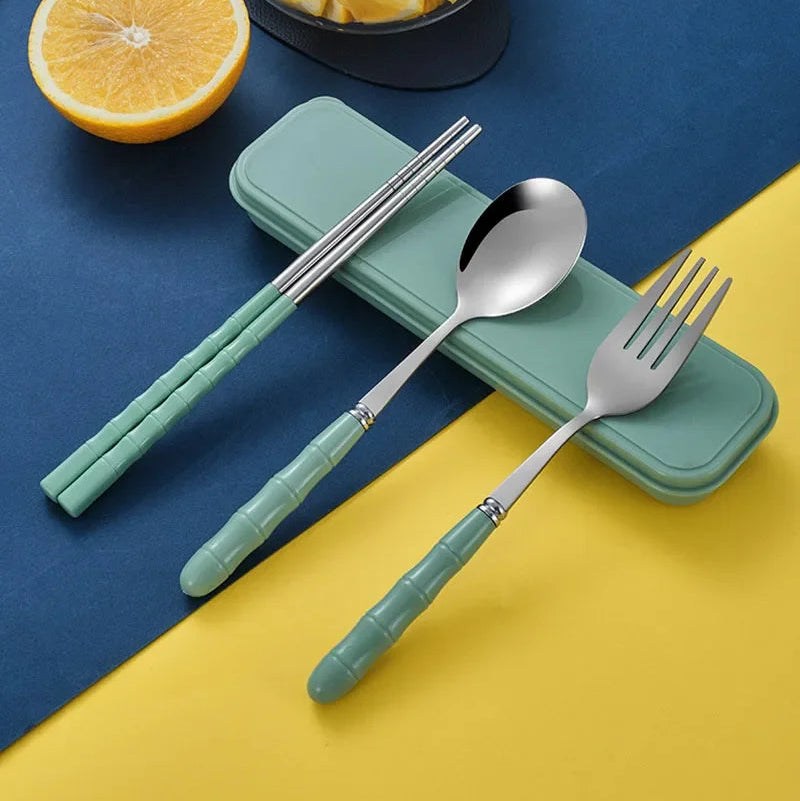 Set of silver kawaii utensils with green handles on a blue and yellow background, next to a half-sliced orange and a bowl of fruit.