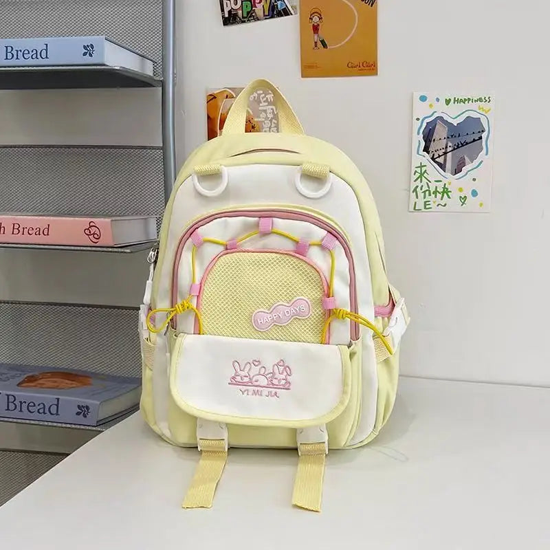 A pastel yellow and white backpack with pink accents, featuring a front mesh pocket with a pink bow and the text 'Happy Birthday', displayed on a white table next to a gray bookshelf.