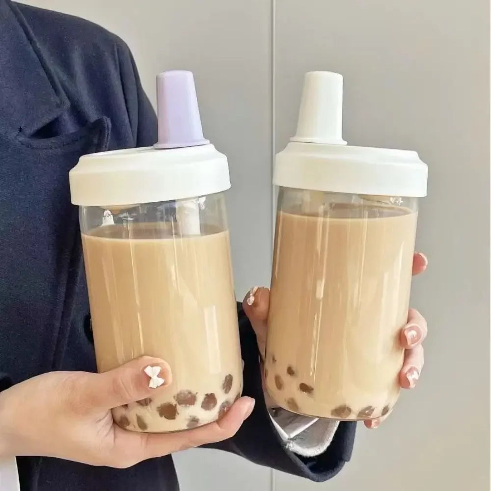 Two hands holding clear kawaii cups with white lids filled with a creamy beverage with visible tea balls.