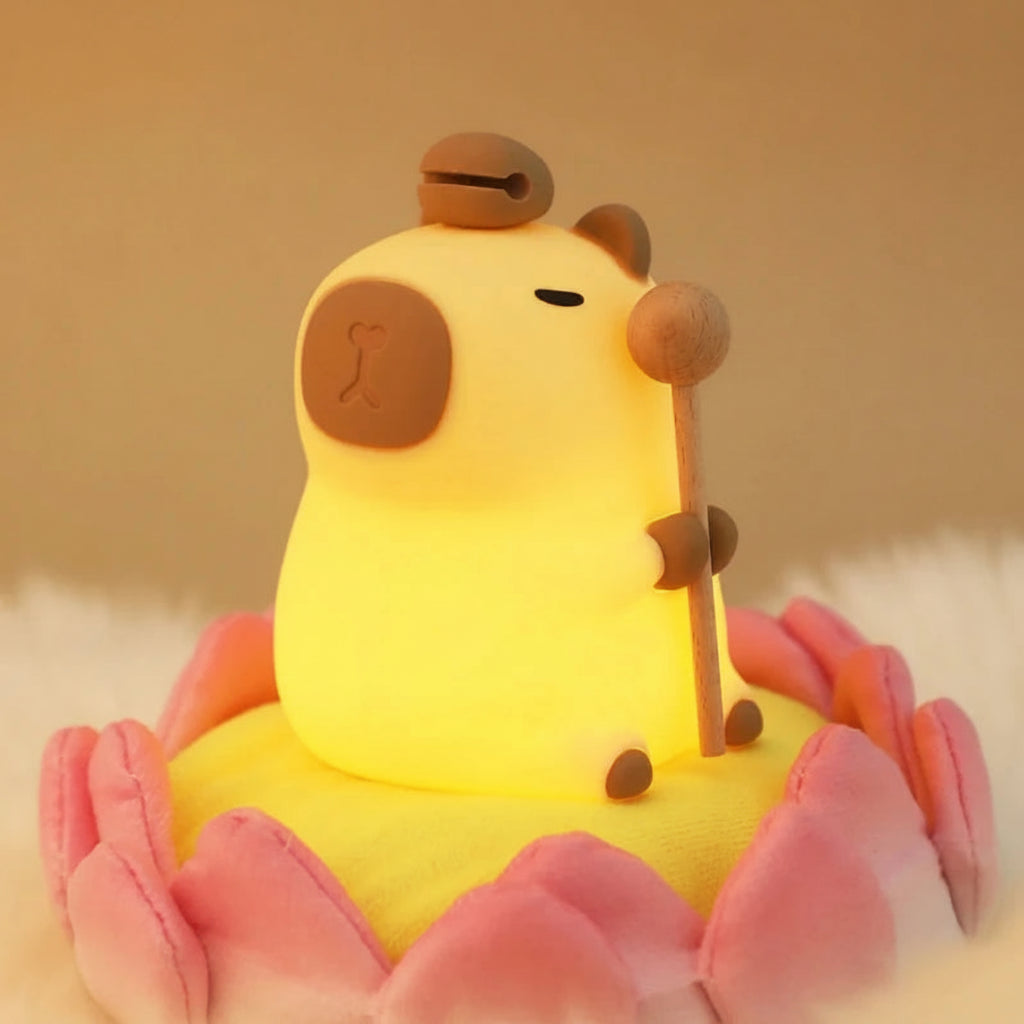 A cute yellow pig-shaped night light with a wooden staff, resting on a pink lotus flower base. The product is illuminated with a warm yellow glow, set against a soft beige background with the 'Twinkle Tots' brand logo in the top-left corner.
