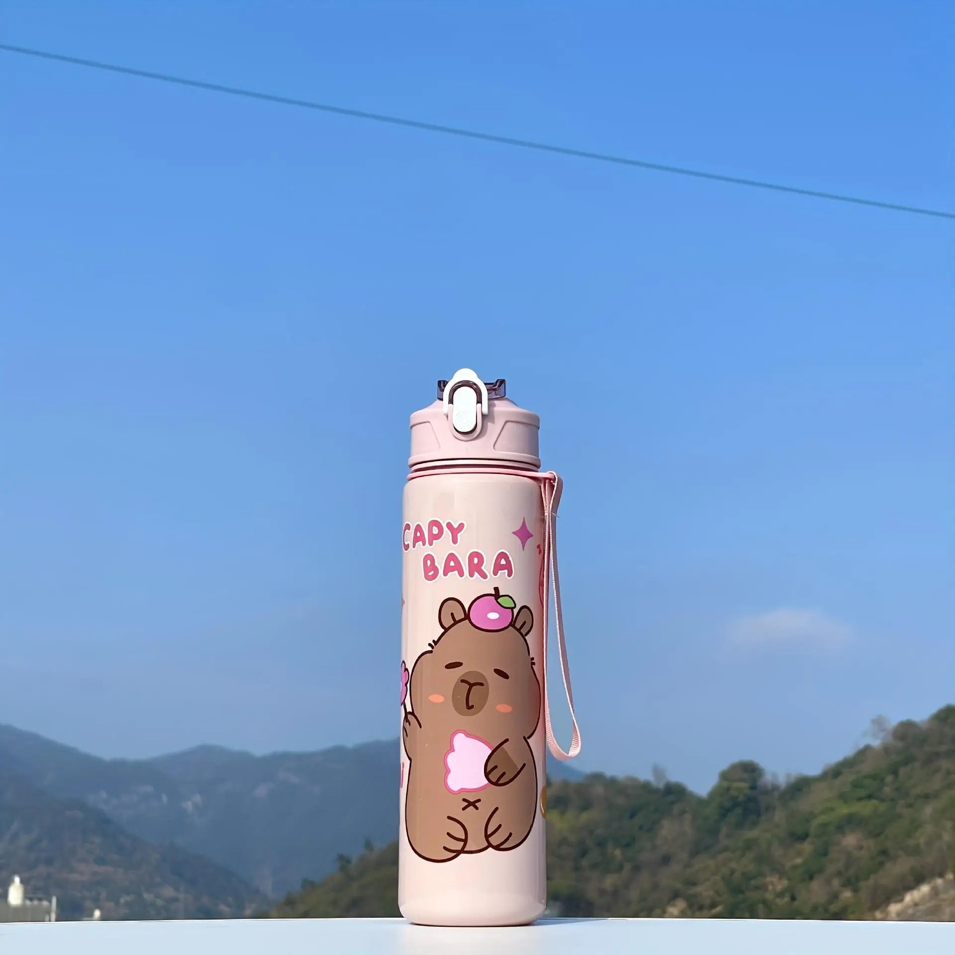 Pink kawaii water bottle with cartoon bear design and 'Capybara' text against a blue sky and mountain background