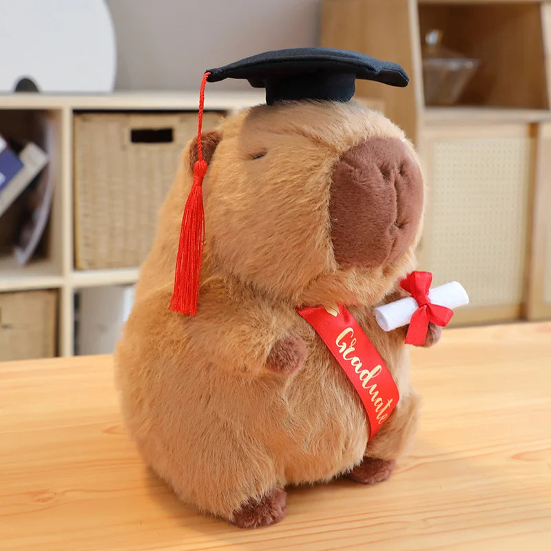Capybara plush toy resembling a groundhog wearing a graduation cap and holding a diploma on a wooden surface.