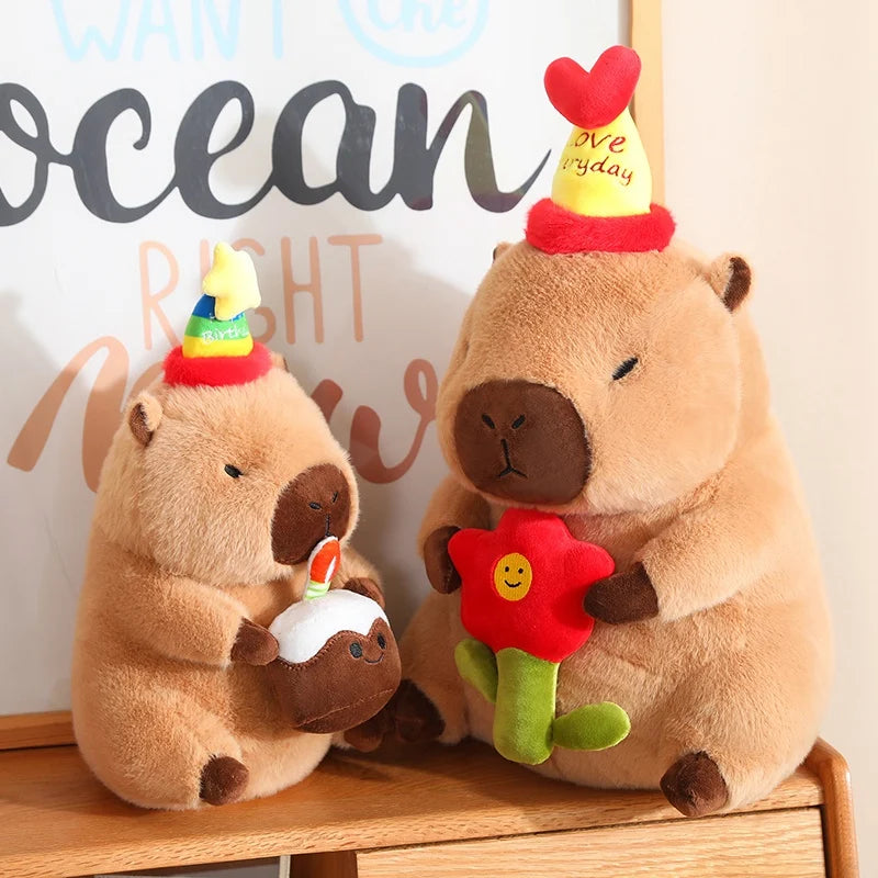 Two capybara plush toys with birthday hats and a flower on a wooden surface.