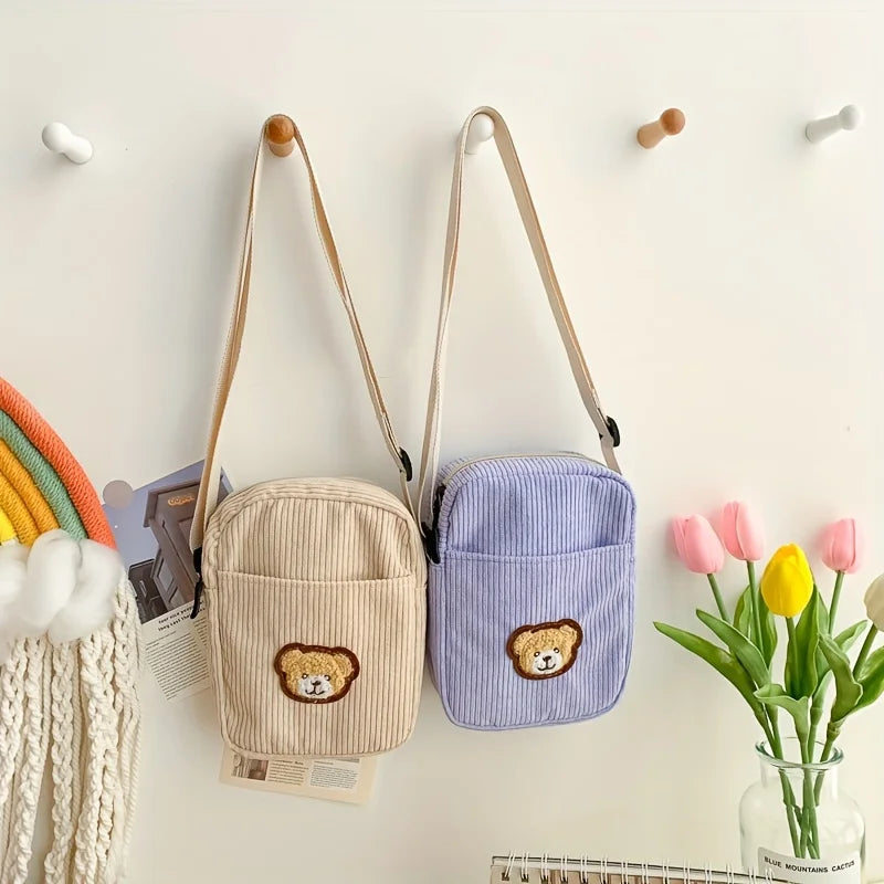 Two small corduroy crossbody bags hanging on pegs against a white wall, one in beige and one in light purple, each with a teddy bear face patch on the front pocket. Pink tulips in a glass vase and a rainbow decoration are visible nearby.