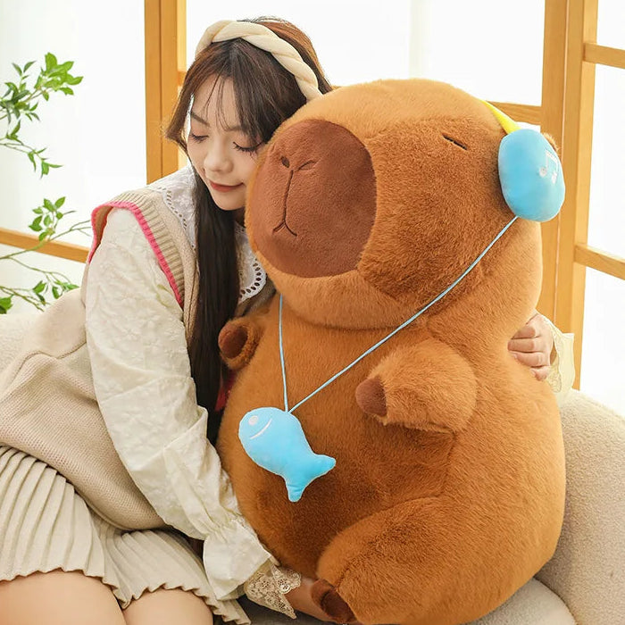 Woman holding a large brown capybara plush toy with headphones in a room with wooden flooring and plants.